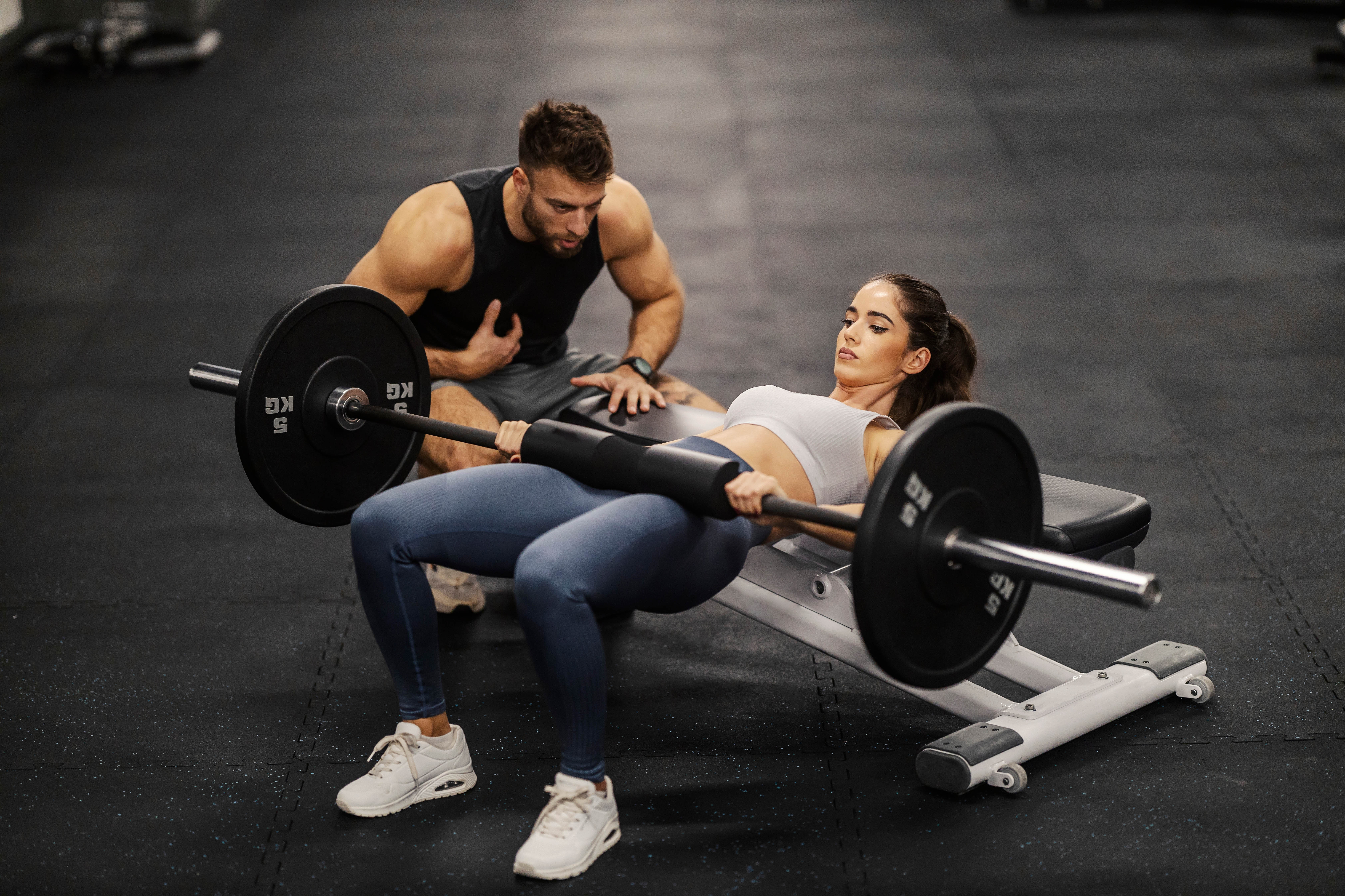 Young female client and male personal trainer doing a hip thruster in the middle of a gym Young female client and male personal trainer doing a hip thruster in the middle of a gym
